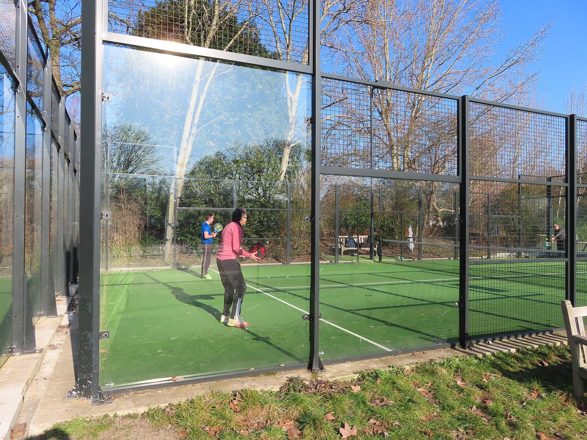 Arka duvarı cam, üstü ve yanları tel örgüyle çevrili açık hava padel kortu; zeminde servis çizgileri görünüyor