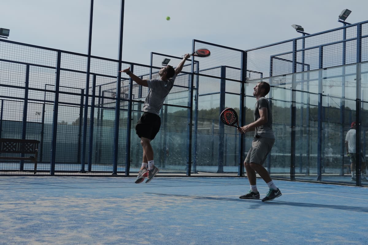 Açık hava padel kortunda iki oyuncu: biri havadaki topa sıçramış, öteki raketi hazırda bekliyor
