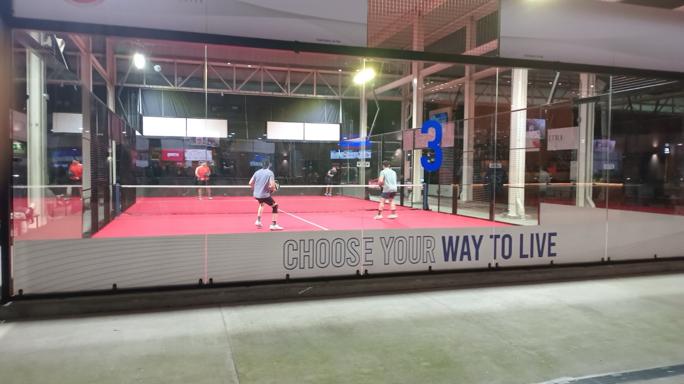 Cam duvarlarla çevrili bir padel kortunda file etrafında oynayan dört oyuncu